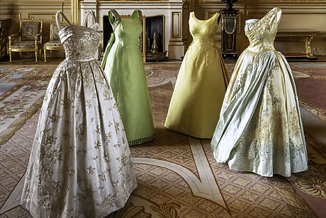 Four elegant vintage evening gowns displayed in a luxurious room with ornate furniture and intricate carpet patterns.