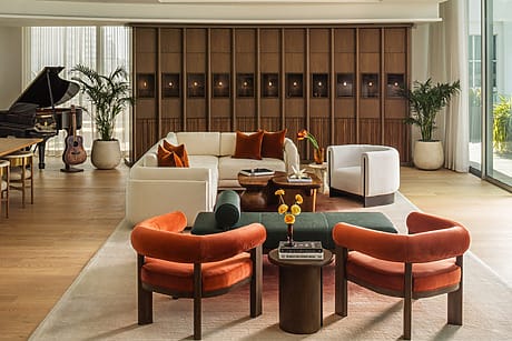 Modern living room with white sectional, orange chairs, wooden accents, plants, and a grand piano in the corner.