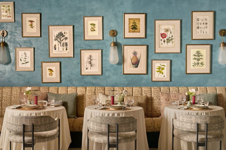 Cozy restaurant interior with teal walls, plant art, round tables with white cloths, and patterned chairs.