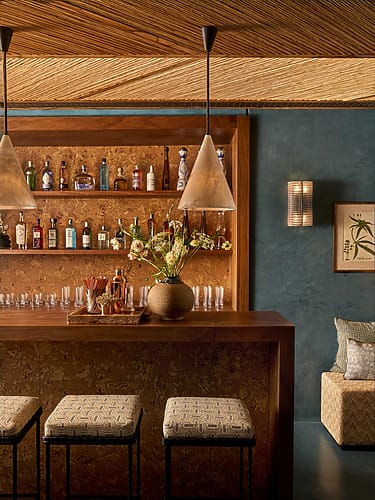 Stylish home bar with wooden counter, stools, hanging lights, and various liquor bottles in a cozy, warmly-lit setting.