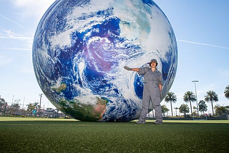 Person in gray coveralls standing next to a giant Earth globe outdoors on a grassy field with palm trees in the background.