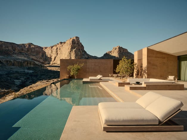 Modern outdoor lounge area with an infinity pool, sunbeds, and a mountainous backdrop under a clear blue sky.