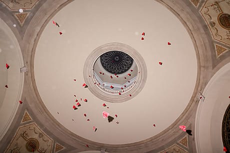 Hearts falling through a circular skylight in a grand architectural ceiling with intricate moldings.