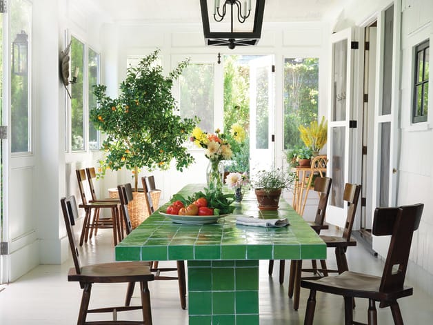 Bright dining room with green tiled table, wooden chairs, and fresh flowers and fruit, surrounded by large windows.