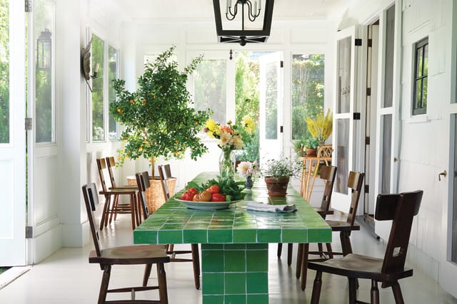 Bright dining room with green tiled table, wooden chairs, and fresh flowers and fruit, surrounded by large windows.