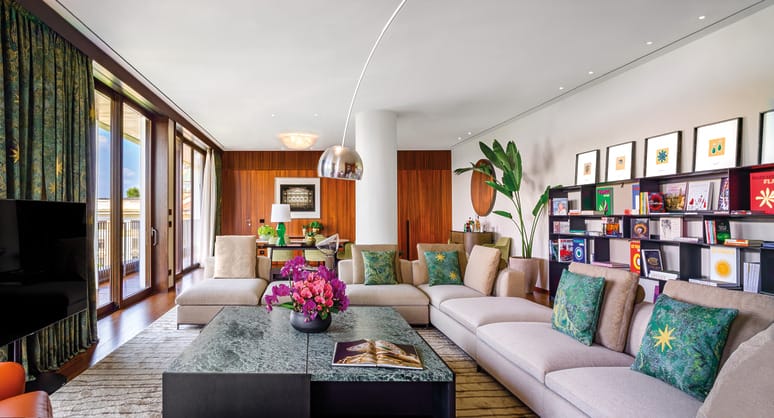 Modern living room with plush sofas, colorful cushions, marble coffee table, bookshelf, and a large plant by floor-to-ceiling windows.