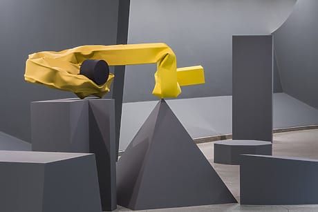 Modern art installation with abstract yellow and black shapes surrounded by geometric gray structures in a gallery setting.