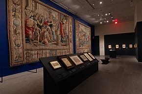 Museum gallery with large, detailed tapestries on blue walls and framed artworks displayed on a dark stand.