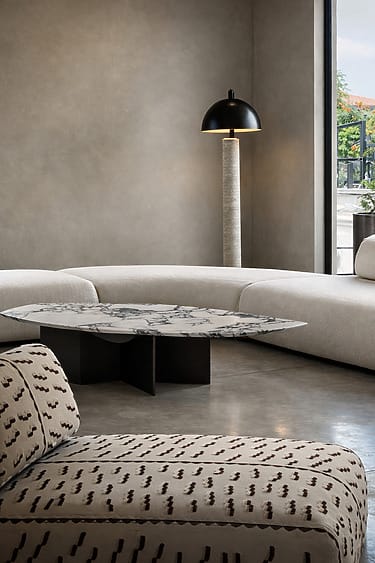Modern living room with curved beige sofa, patterned armchair, marble coffee table, and standing lamp.