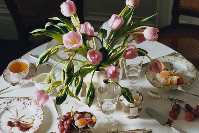 Table set for breakfast with pink tulips, tea, grapes, cherries, and bread on patterned china plates.