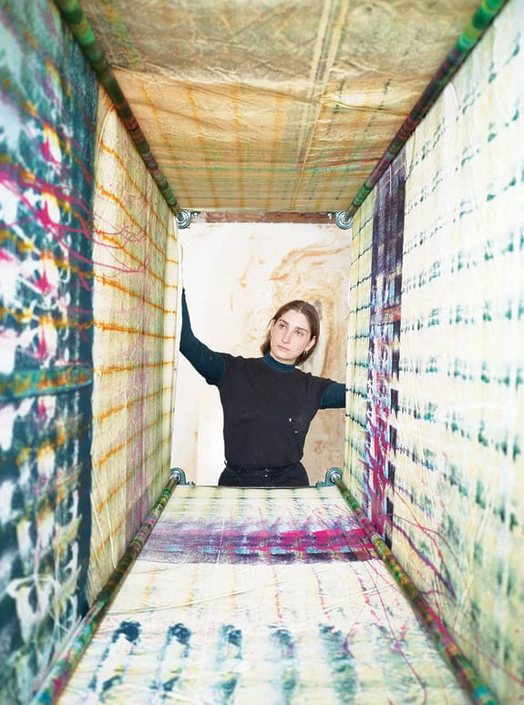 Person standing inside colorful, patterned textile structure, looking outwards, surrounded by abstract design elements.