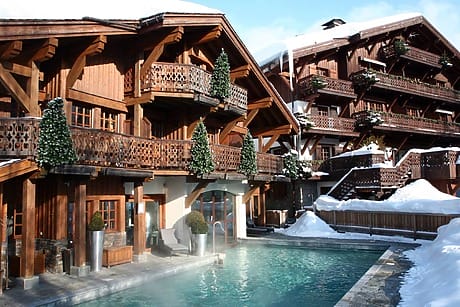 Mountain chalet with wooden balconies, snowy surroundings, and a steaming outdoor pool, framed by evergreen trees.