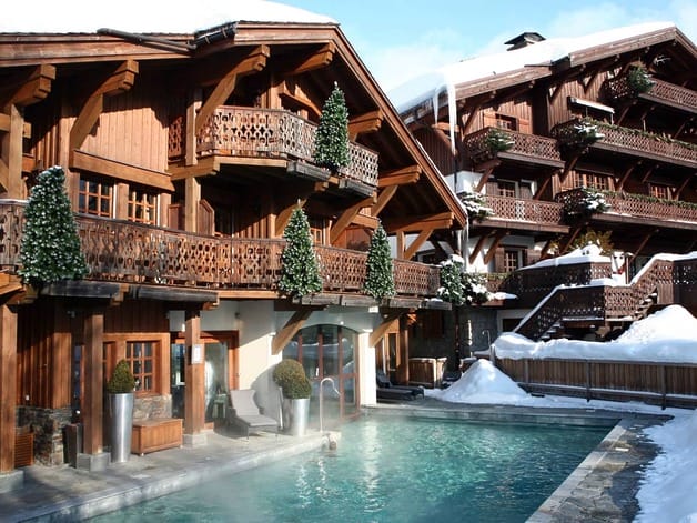 Mountain chalet with wooden balconies, snowy surroundings, and a steaming outdoor pool, framed by evergreen trees.