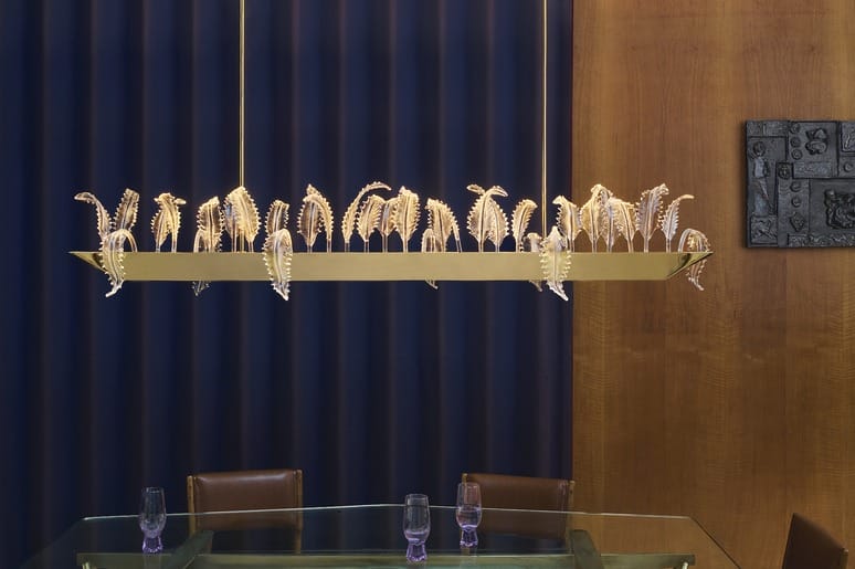 Elegant chandelier with intricate leaf design hanging over a glass dining table, set against a navy curtain background.