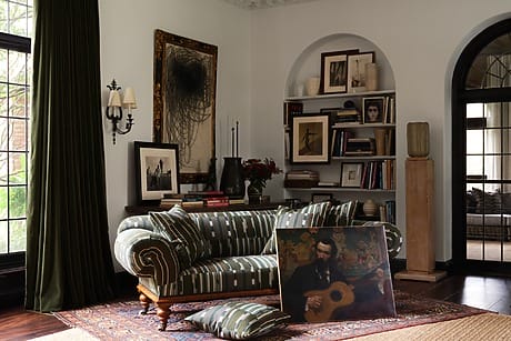 Elegant living room with a striped sofa, scattered art pieces, a bookshelf, and large windows with dark drapes.
