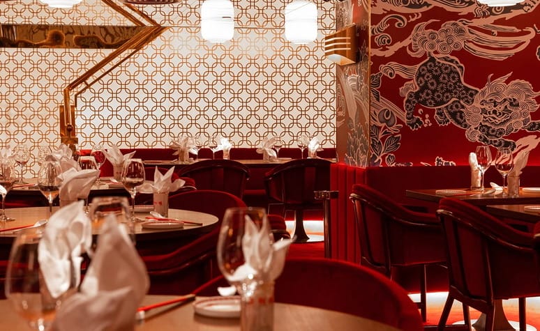 Elegant restaurant interior with red and gold decor, intricate wall patterns, and tables set with white linens and glassware.