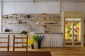 Cozy kitchen with open shelves, flowers, and a lit "House of Three" fridge display on a wooden floor.