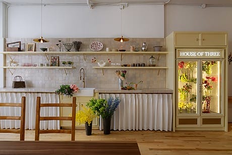 Cozy kitchen with open shelves, flowers, and a lit "House of Three" fridge display on a wooden floor.