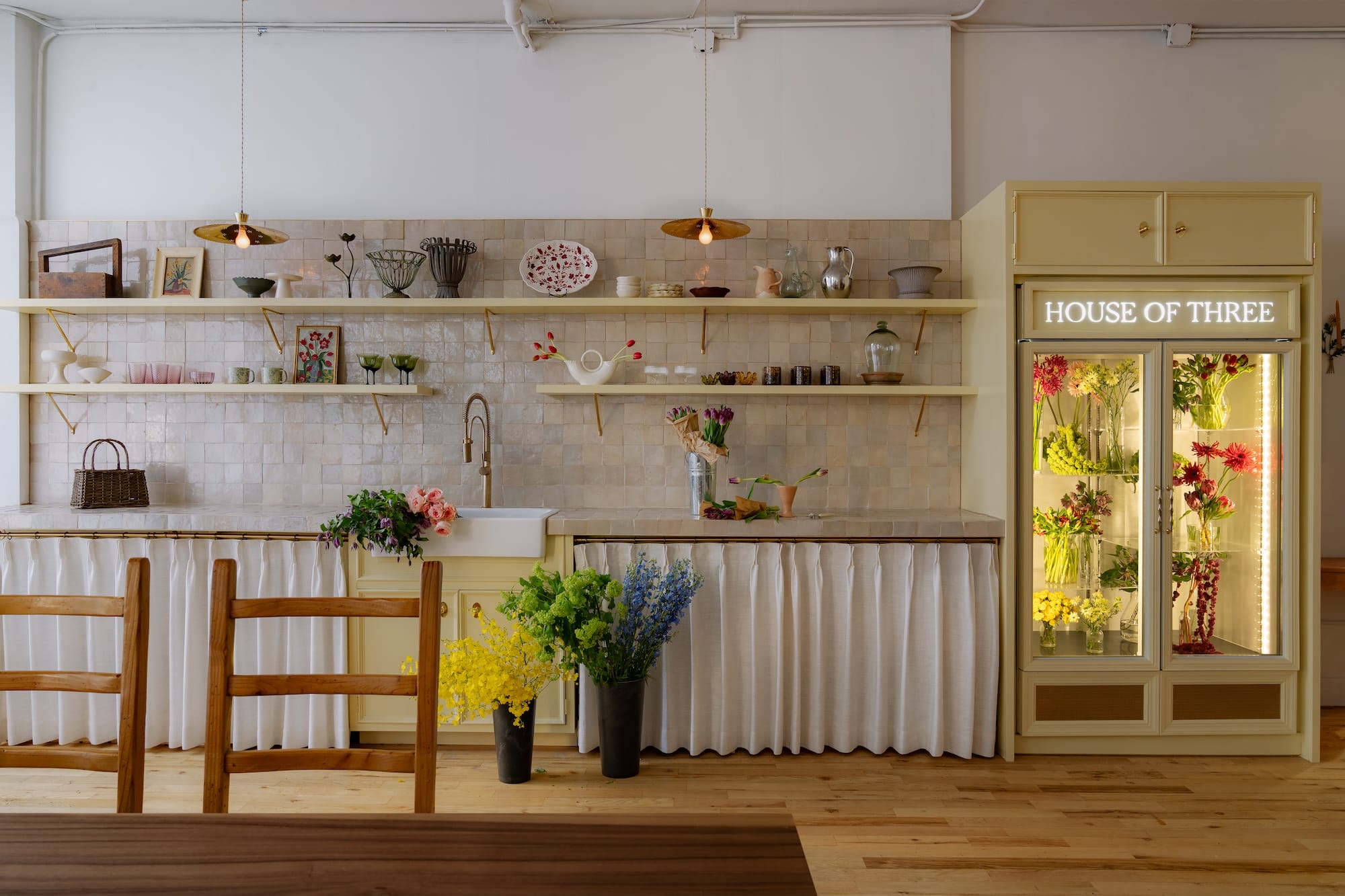 Cozy kitchen with open shelves, flowers, and a lit "House of Three" fridge display on a wooden floor.