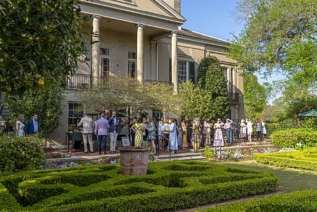 People gathered outside a historic building with columns surrounded by lush gardens on a sunny day.