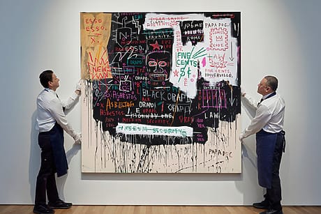 Two men in aprons adjusting a large abstract painting with various words, numbers, and symbols on a white gallery wall.