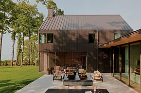 Modern house with dark wood exterior, large windows, and patio furniture on spacious lawn surrounded by tall trees.
