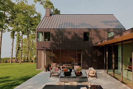 Modern house with dark wood exterior, large windows, and patio furniture on spacious lawn surrounded by tall trees.
