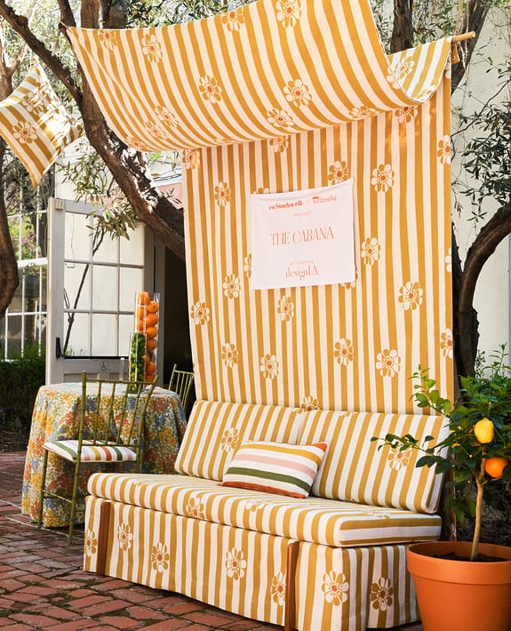 Outdoor patio with yellow striped and floral cushions, canopy, and curtains, next to a table with a matching tablecloth.