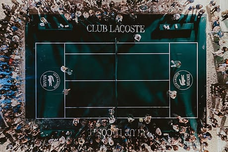 Aerial view of a crowded tennis court at Club Lacoste with people gathered around the edges.