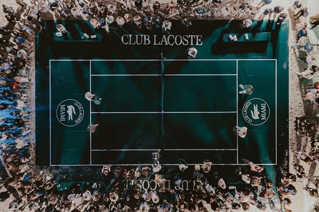 Aerial view of a crowded tennis court at Club Lacoste with people gathered around the edges.