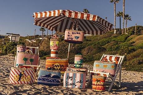 Colorful beach bags displayed under a striped umbrella on sandy beach with palm trees and greenery in the background.