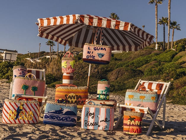 Colorful beach bags displayed under a striped umbrella on sandy beach with palm trees and greenery in the background.