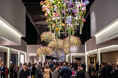 People gathering at a vibrant art gallery event with colorful floral displays hanging from the ceiling.