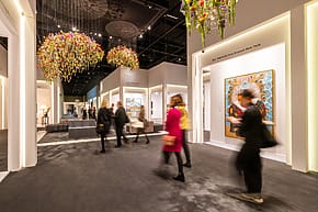 Visitors walking through a modern art gallery with colorful chandeliers and large paintings in a well-lit exhibition space.