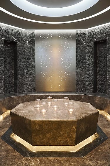 Luxurious spa room with a hexagonal stone table, ambient lighting, candles, and a decorative wall panel with circular patterns.