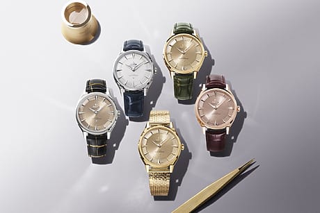 Five luxury watches with different straps and colors, displayed on a table with a gold ring and tweezers nearby.