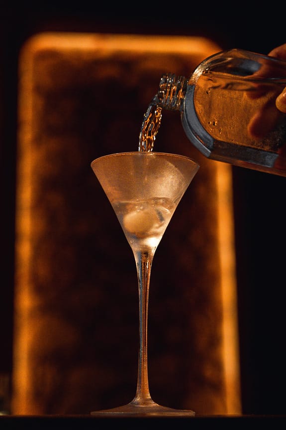 Martini glass with ice being filled with liquid from a bottle against a warm, glowing background.