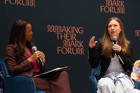 Two women engaged in discussion on stage at the Making Their Mark Forum, with a microphone and a dark background.