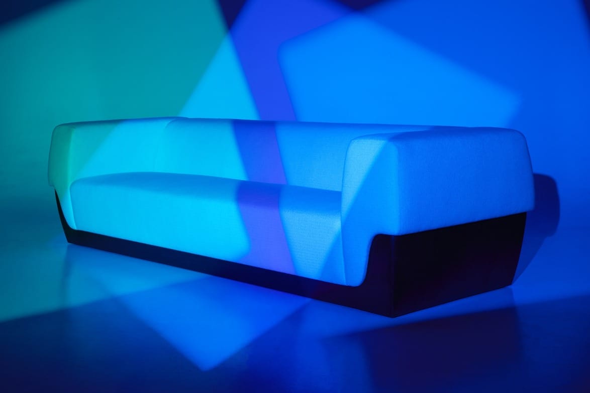 Modern blue and cyan designer sofa illuminated with colorful geometric light patterns in a minimalist setting.