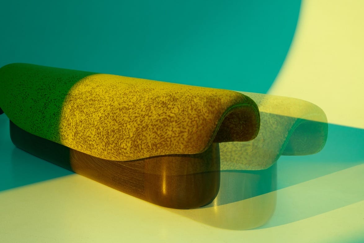 Modern bench with a yellow speckled cushion and wooden base, casting a shadow under vibrant green and yellow lighting.