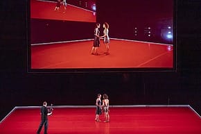 Two dancers performing on a red stage with a large screen displaying the same scene above them.