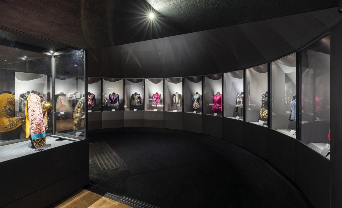 Installation view of fashion exhibit on Elsa Schiaparelli at the Victoria and Albert Museum in London displaying a collection of colorful, ornate jackets in a dimly lit, curved glass case.