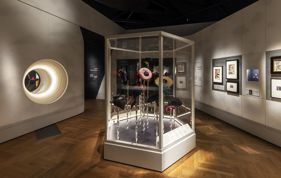 Installation view of fashion exhibit on Elsa Schiaparelli at the Victoria and Albert Museum in London with art museum gallery with modern sculptures in a glass display case and framed artworks on walls.