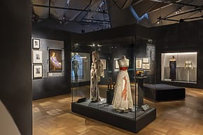 Installation view of fashion exhibit on Elsa Schiaparelli at the Victoria and Albert Museum in Londonfeaturing historical costumes in a dimly lit room, with framed artwork displayed on the walls.
