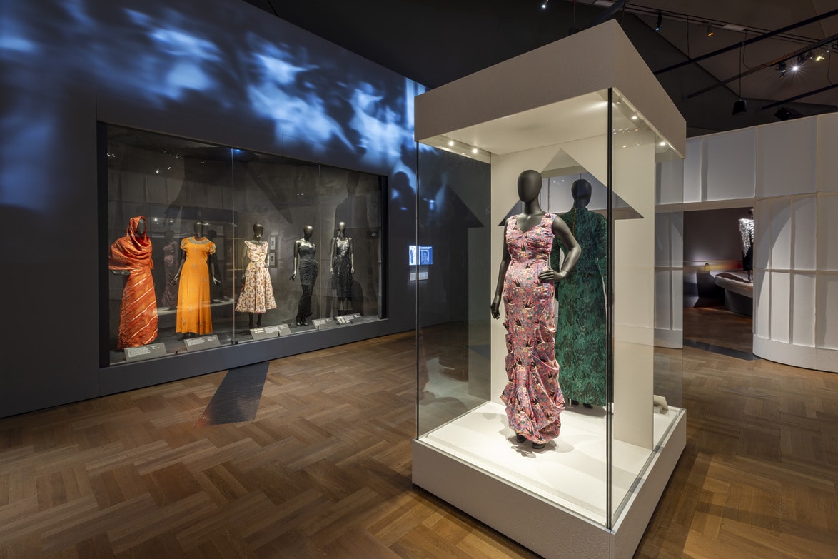 Installation view of fashion exhibit on Elsa Schiaparelli at the Victoria and Albert Museum in London, showcasing colorful vintage dresses on mannequins in a museum setting with wooden flooring and dramatic lighting.