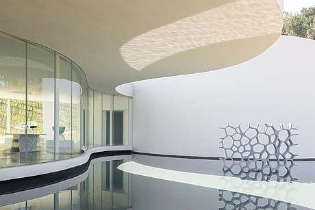 Modern architectural design with curved glass walls and a honeycomb-like sculpture by a reflective water surface.