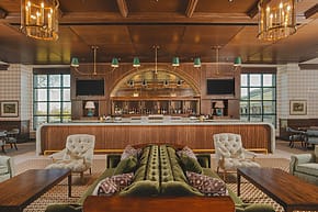 Luxurious hotel lounge with a central bar, green velvet sofas, elegant armchairs, and warm wooden decor.