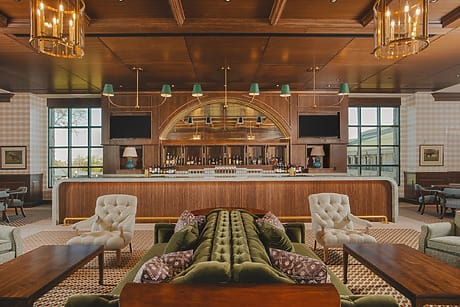 Luxurious hotel lounge with a central bar, green velvet sofas, elegant armchairs, and warm wooden decor.