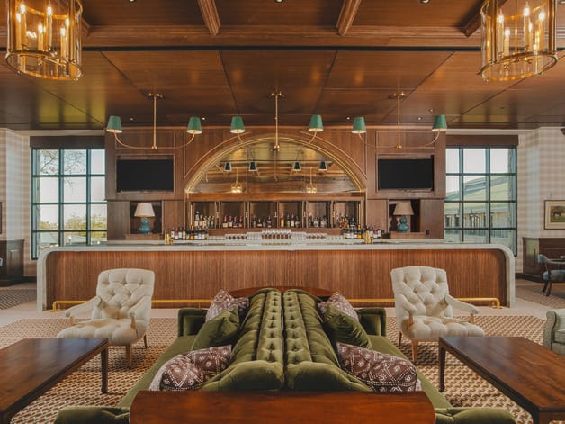 Luxurious hotel lounge with a central bar, green velvet sofas, elegant armchairs, and warm wooden decor.