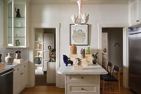 Modern kitchen with marble island, decorative vases, wooden floor, wall art, and glass cabinets.